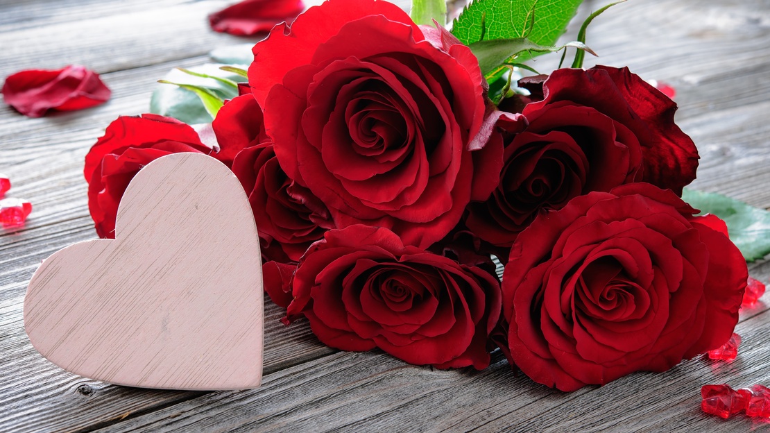 Nahaufnahme übereinander liegender roter Blumen auf einem hölzernen Untergrund. Links vor den roten Blüten steht ein hellrosa Herz aus Holz. Um die Blumen liegen teilweise rote Blütenblätter
