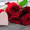 Nahaufnahme übereinander liegender roter Blumen auf einem hölzernen Untergrund. Links vor den roten Blüten steht ein hellrosa Herz aus Holz. Um die Blumen liegen teilweise rote Blütenblätter